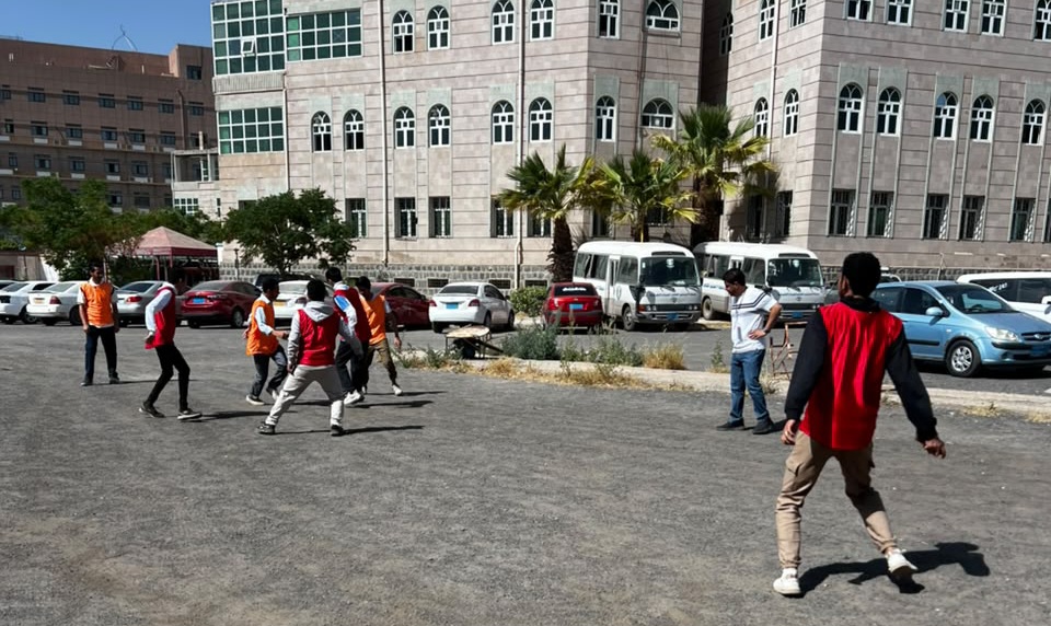 Group of people playing casual soccer in a paved parking lot next to a large campus-style building, several wearing orange or red vests with parked cars in the background and palm trees nearby.