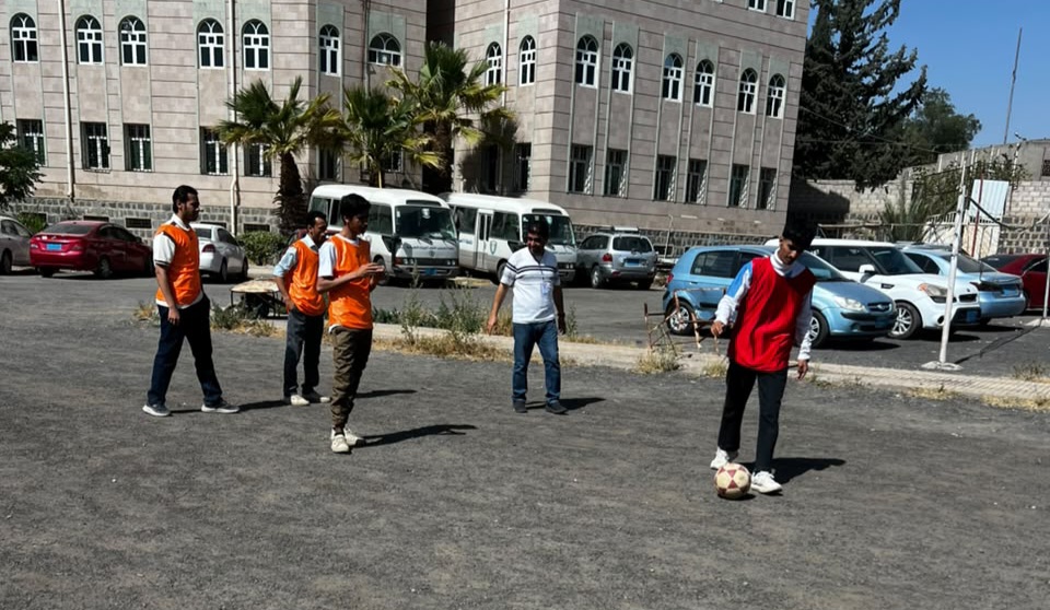 Men in orange vests watching and playing soccer on a paved lot in front of a large building; one player with a red vest controls a soccer ball.