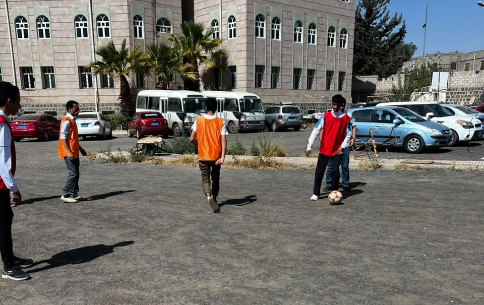 Group of kids playing soccer on a dusty parking lot beside a brick building; a boy in a red bib dribbles the ball while others stand nearby.