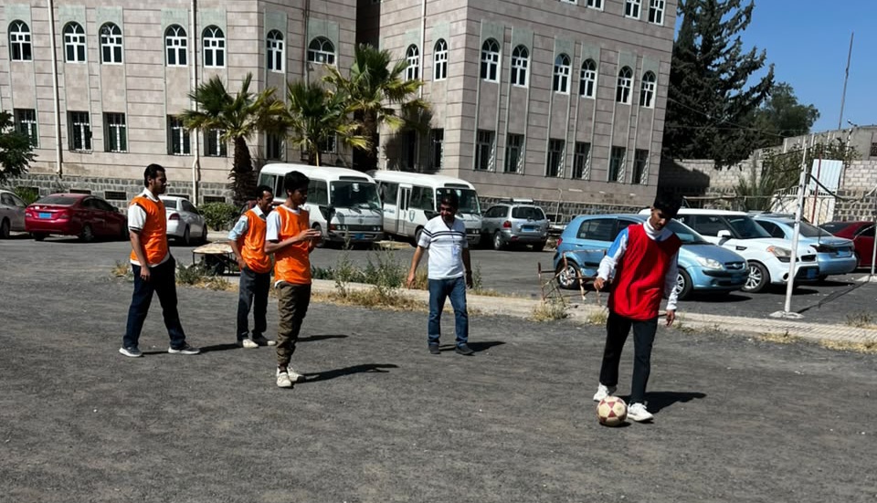 Group of young men playing soccer in a paved lot near a building, one kicking a ball while others watch. Orange vests indicate teams or volunteers.