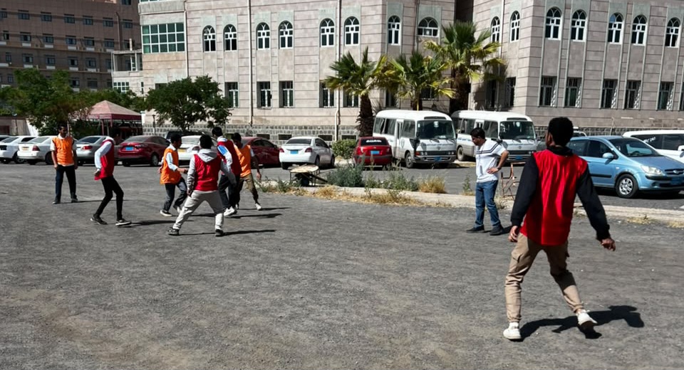Group of people playing soccer in a parking lot beside a campus-style building, wearing orange bibs.