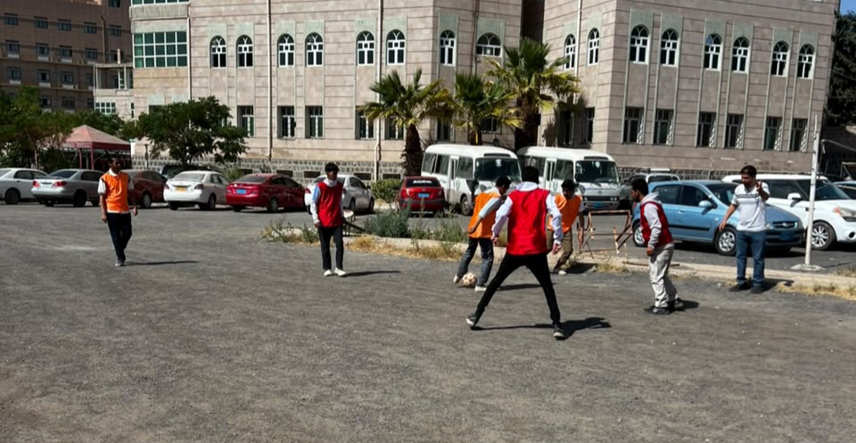 Group of people playing soccer on an asphalt lot in front of a beige campus building; some players wear orange bibs.