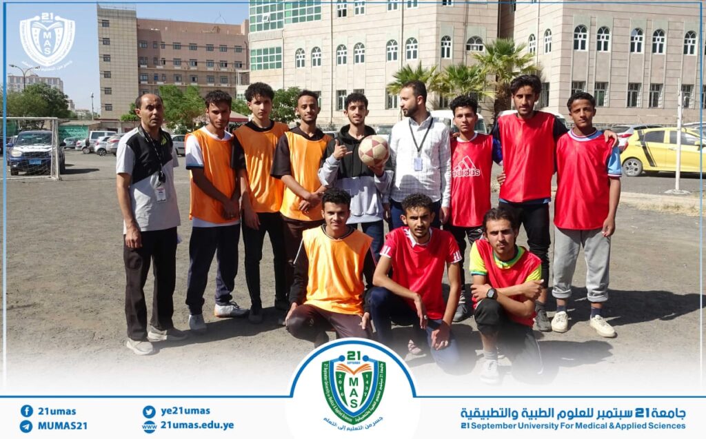 Group of young men in orange and red training bibs posing for a team photo on a dusty outdoor field, with campus buildings in the background and one player holding a soccer ball.