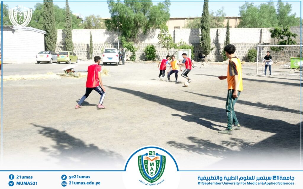 Students playing soccer on a dusty outdoor field at 21 September University for Medical and Applied Sciences, with a goal and trees in the background.