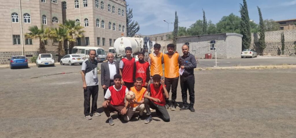 Group of young men posing with a soccer ball on a dusty schoolyard, several wearing red or orange bibs, with a campus building and parked cars in the background.