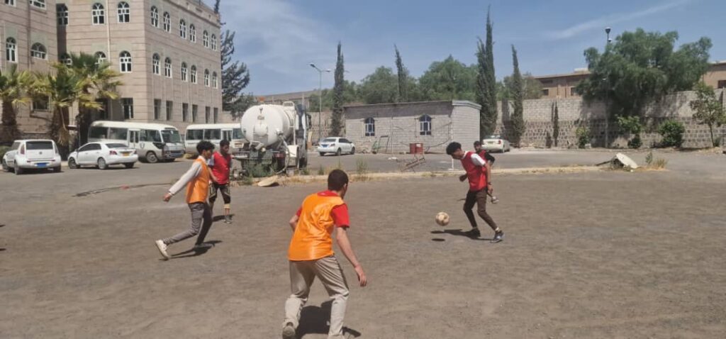 Four young boys wearing orange vests play soccer on a dusty schoolyard with a ball near a beige brick building and parked vans in the background.