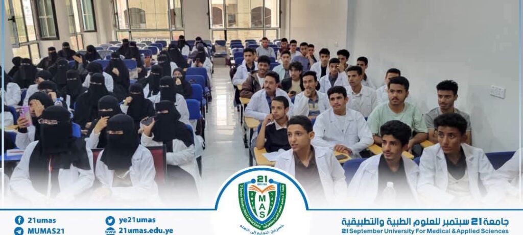 Lecture hall full of students listening to a lecture; several women wear niqab in front rows, some students in white coats, with university banner/logo at the bottom.