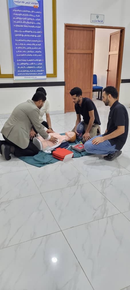 Three men in a hallway practice CPR on a medical dummy placed on a mat; one performs chest compressions while two observe nearby.