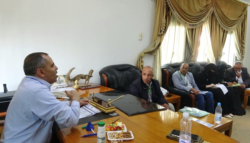 Professionals seated around a large conference table, documents and snacks laid out, in a formal meeting room with gold curtains.