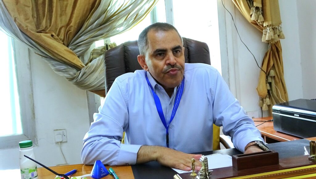 Man in a light blue shirt sits at a desk in an office with papers, pens, and a printer nearby, wearing a blue lanyard.