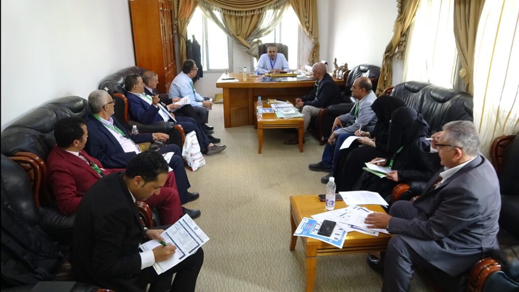 A formal meeting in a sunlit office with people seated along leather couches around a central wooden desk, papers and notebooks in hand as they discuss.
