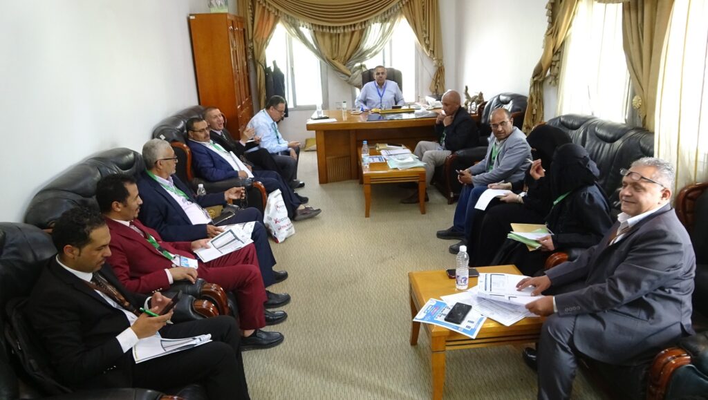 Group of professionals seated along leather couches around a central desk, discussing with papers and water bottles on tables in a formal office.