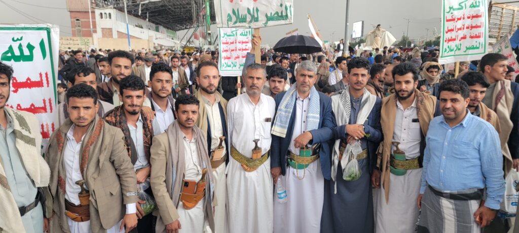 Crowd of men in traditional Yemeni attire at a march, many with daggers at their belts, holding Arabic banners.