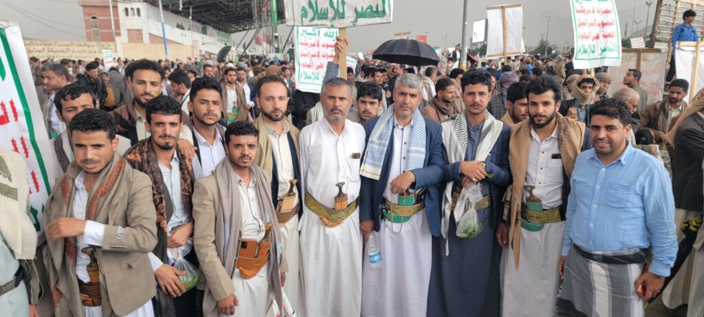 A large group of men in traditional Yemeni clothing gathered for a street protest, many wearing scarves and swords; signs with Arabic text are visible above them.