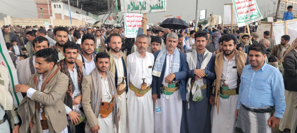 Large crowd of men in traditional Yemeni dress at a street demonstration, holding Arabic banners; many wear scarves and ceremonial daggers.