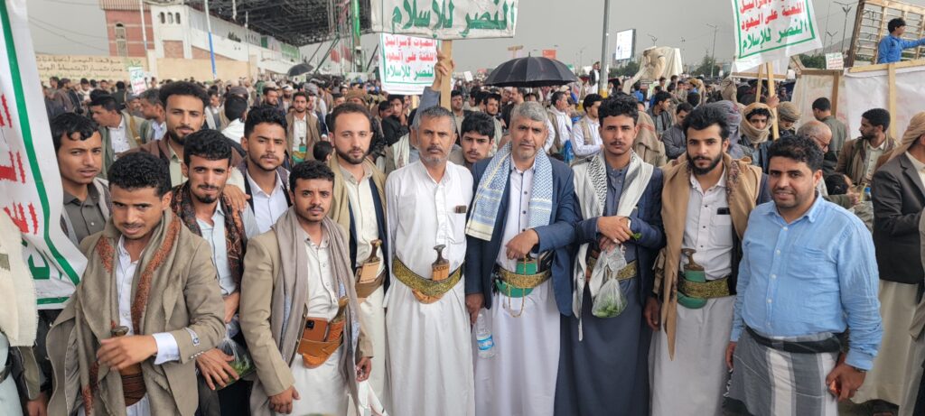 Large group of men in white thawbs and beige jackets at a street rally, banners in Arabic overheads.