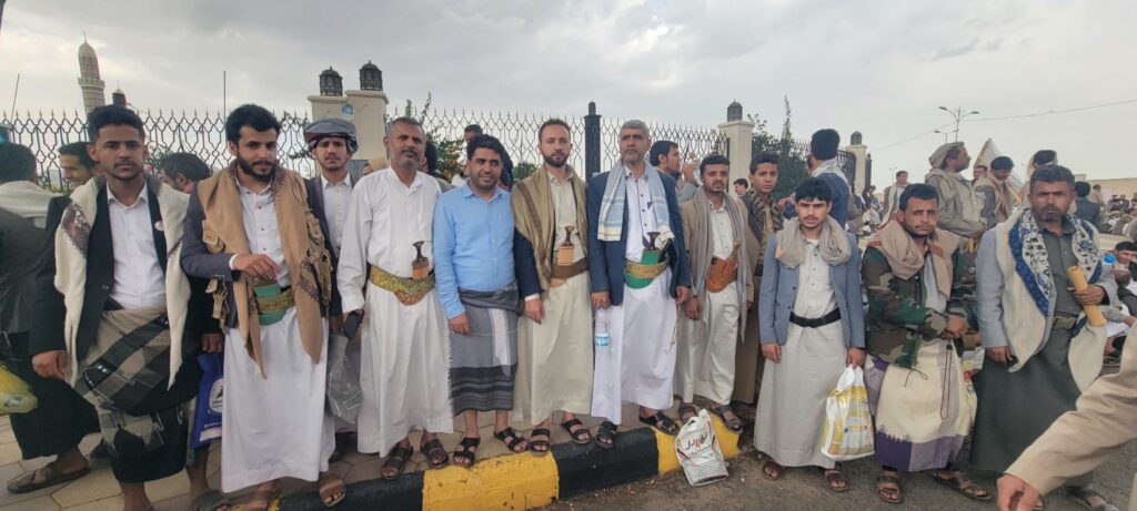 Group of men in traditional robes and scarves stand in a line along a decorative fence, some holding bags and sodal items, on a cloudy day.