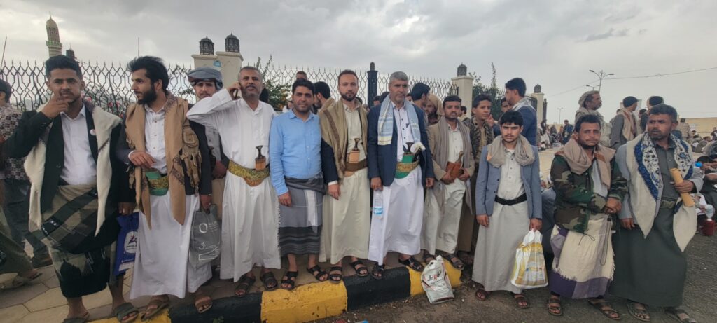 Group of men in traditional Yemeni dress standing along a curb beside a decorative fence, some with swords and bags, with a cloudy sky behind them.