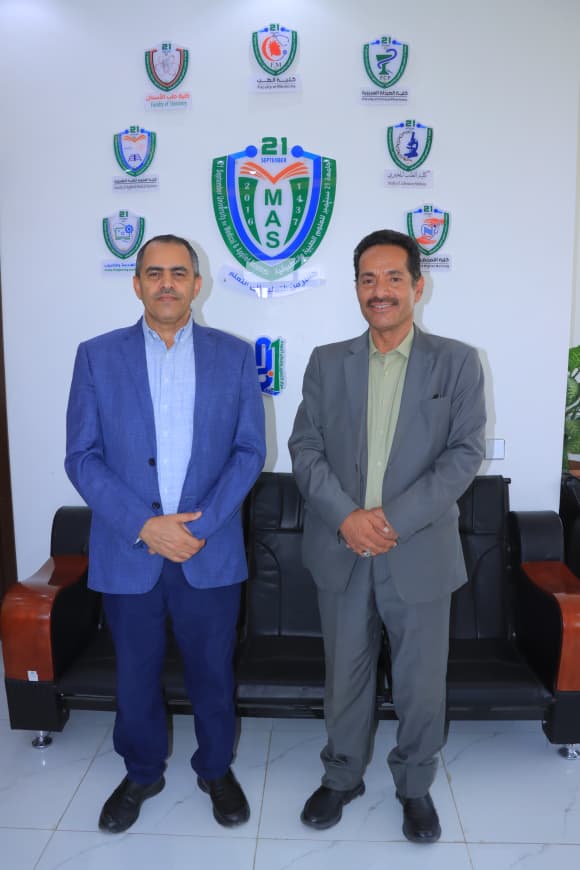 Two men in suits pose in front of a wall with multiple blue‑green emblems and a large central MAS shield logo behind them.