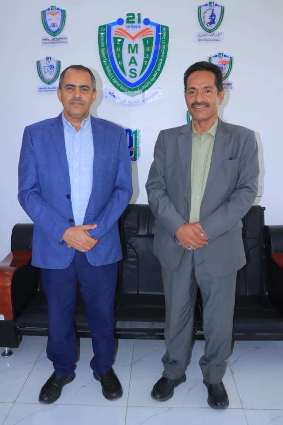 Two men in business suits stand side by side in front of a wall with MAS emblems in the background, posing for a photo.