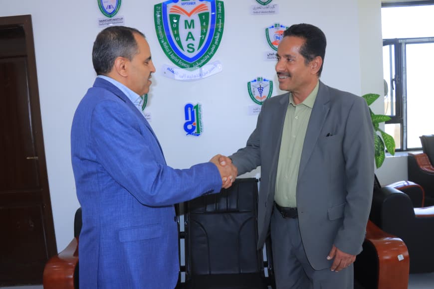 Two men in suits shake hands in an office with multiple organizational logos on the wall behind them.