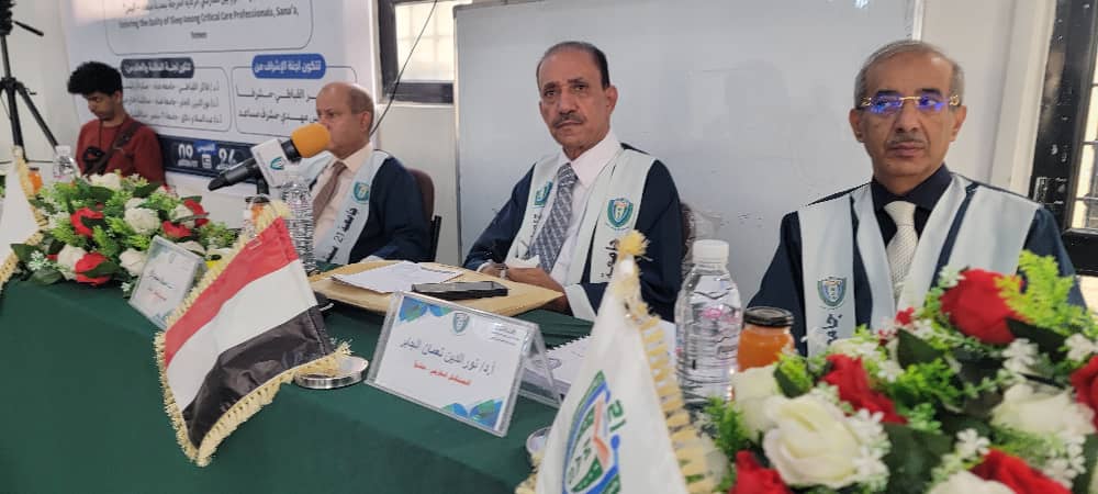 Panel of three men in vests seated at a green-draped table with microphones, flags, and floral arrangements at a formal conference (Iraqi flag visible).