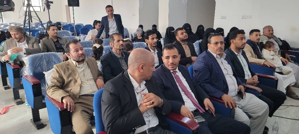 Audience seated in a conference hall, mostly men in suits, with women in hijabs visible in the back rows.