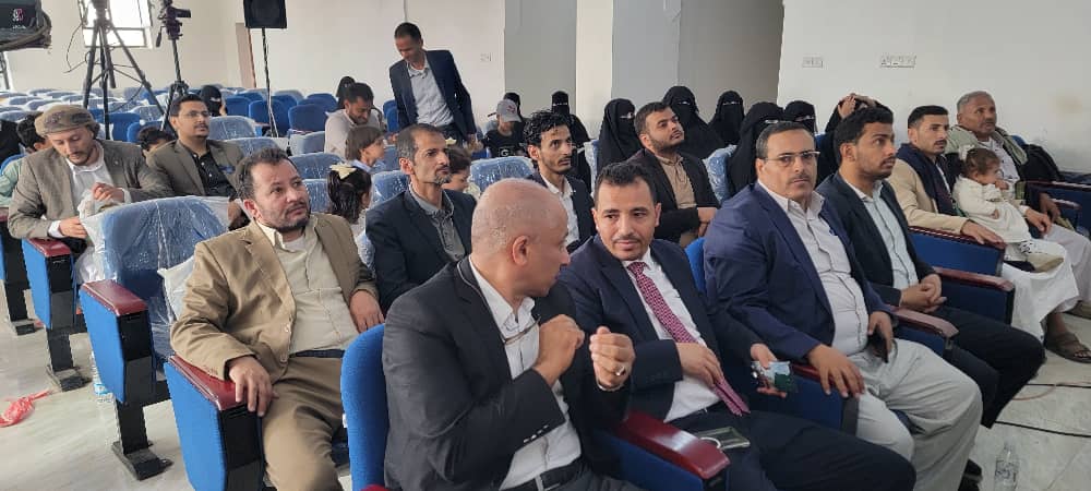 Audience in a lecture hall: men in suits seated in front rows, chatting, with women in black hijabs in back rows.
