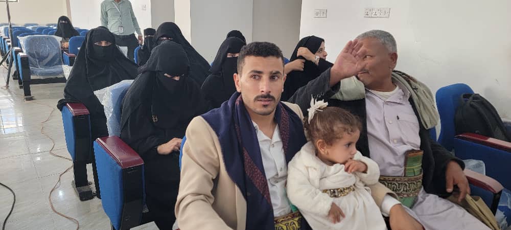 Man in a beige coat sits in a clinic waiting area holding a baby girl in white; an older man beside him rests his hand near his face while veiled women sit in the background.