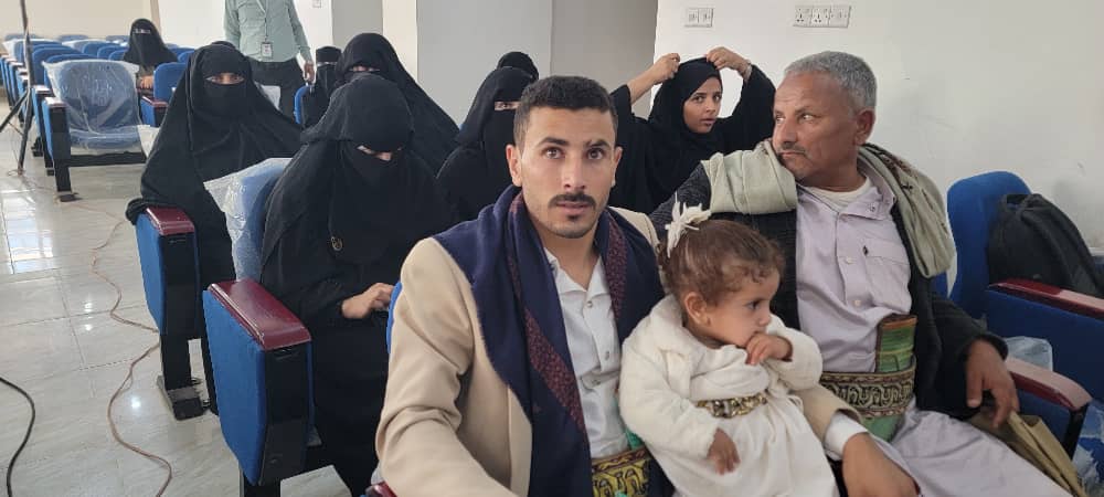 Man with a mustache in a tan suit holds a young girl in a white dress while seated in a waiting room; several women in black hijabs and an older man sit nearby.