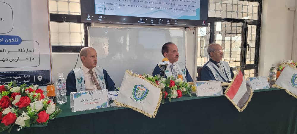 Three men in suits sit at a long conference table on a panel, with flag banners, nameplates, and floral arrangements in front, during a formal meeting offering a presentation on a screen above them.