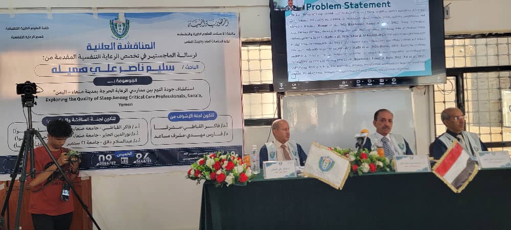 Panel discussion at a conference with three speakers seated behind a long table, flags and flower arrangements, and a large Arabic banner on the wall; a monitor above shows a slide titled'Problem Statement'.