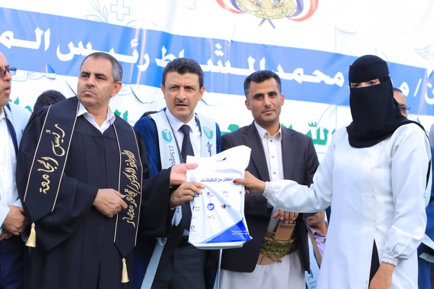1003982185 - جامعة 21 سبتمبر Group of officials on a stage handing a bag to a woman in a niqab during a formal ceremony; blue Arabic banner in the background revealing organizational context.