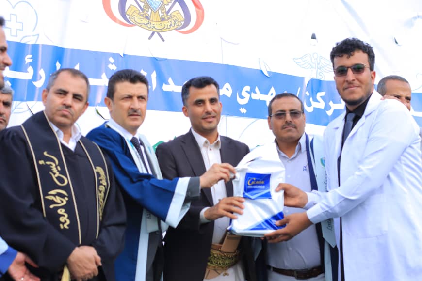 1003982188 - جامعة 21 سبتمبر Group of men in ceremonial robes and suits present a boxed item to a man in a white coat at an outdoor event with a blue banner in the background.