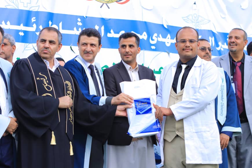 1003982191 - جامعة 21 سبتمبر Group of men on a stage; a man in a white coat hands a blue-and-white bag to a recipient in a suit, with a blue Arabic banner in the background.