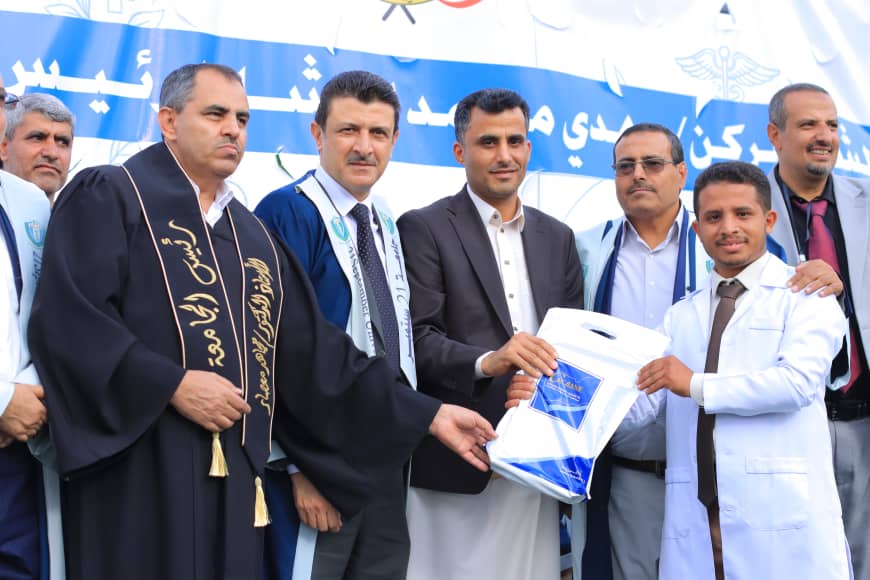 1003982194 - جامعة 21 سبتمبر Group of formally dressed men on a stage during a ceremony, one man in dark robe hands a package to a man in a white coat while others look on.