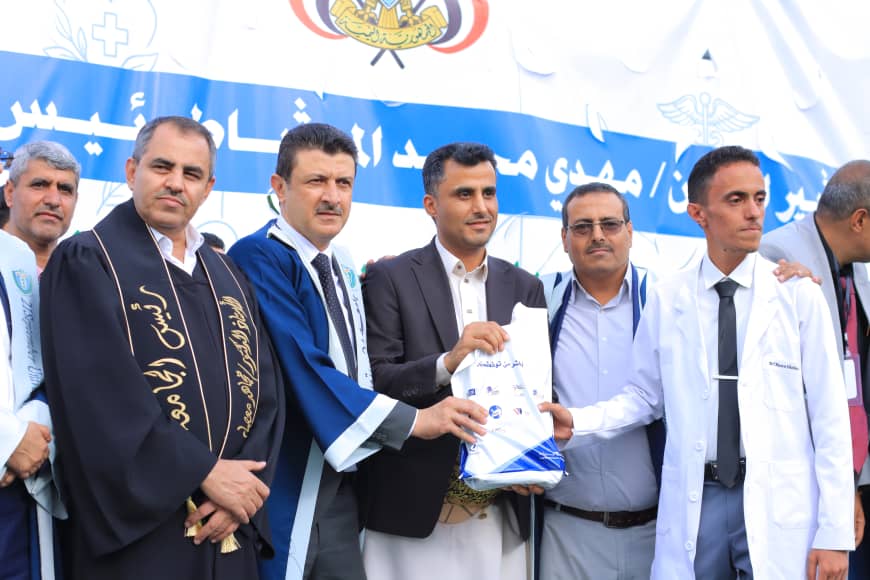 1003982197 - جامعة 21 سبتمبر Group of men in formal attire on stage during an award ceremony, one handing a packet to another amid a blue banner backdrop