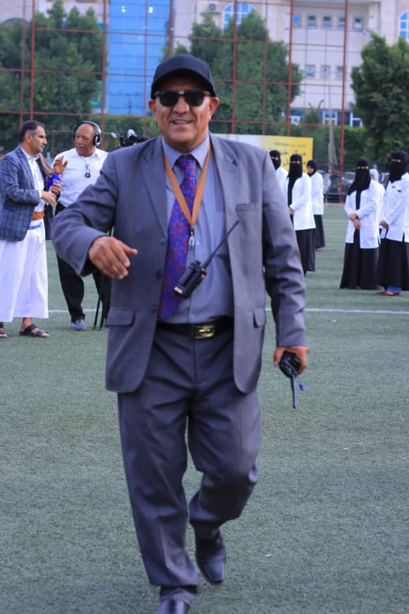 1003982200 - جامعة 21 سبتمبر Smiling man in a gray suit and cap walks toward the camera on a sports field, wearing sunglasses and a lanyard with a radio, with people in white robes behind him.