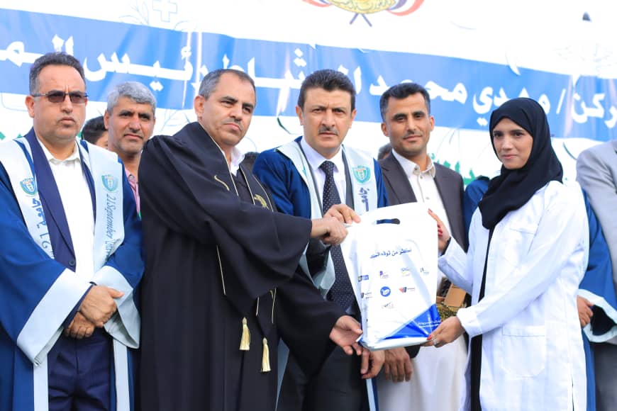 1003982203 - جامعة 21 سبتمبر Group of men in robes presents a white gift bag to a woman in a lab coat at an outdoor ceremony, blue banner in background with logos nearby.