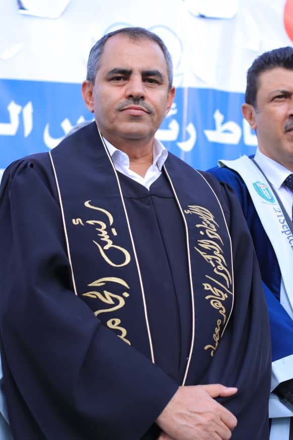 1003982206 - جامعة 21 سبتمبر Man in a dark ceremonial gown with gold embroidery stands at an outdoor event, with a blue banner behind and a second man beside him.
