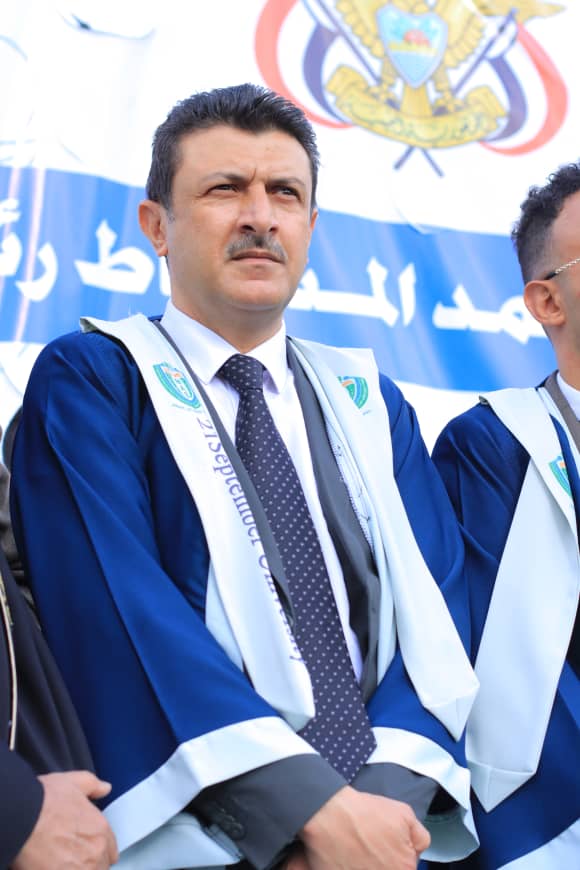 1003982212 - جامعة 21 سبتمبر Man in a blue ceremonial uniform and dotted tie, standing at a formal event, with a banner and crest visible in the background.