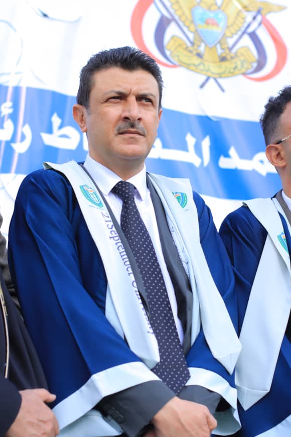 1003982215 - جامعة 21 سبتمبر Man in a blue academic gown and tie sits at a ceremony, with a banner and crest in the background.