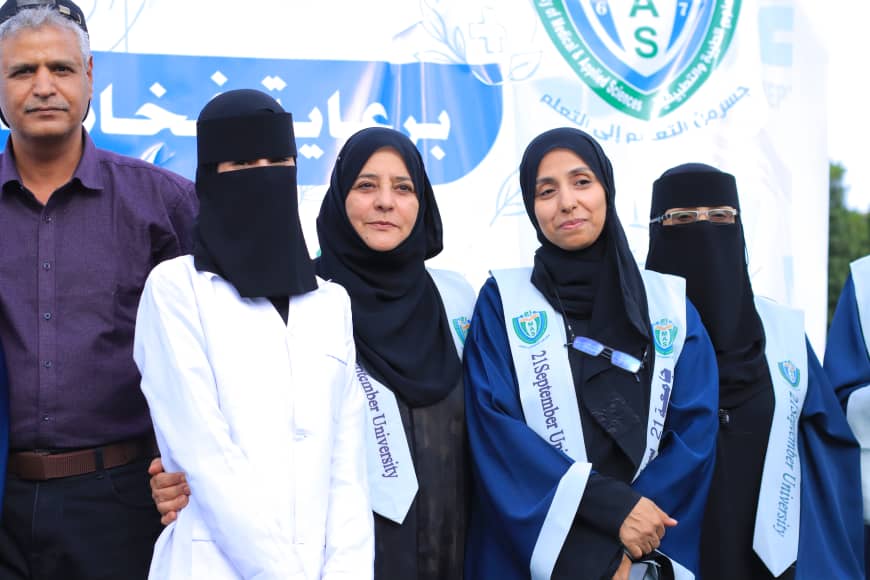 1003982218 - جامعة 21 سبتمبر Group of women in hijabs and sashes at a university graduation event, posing in front of a banner with a crest logo and text in Arabic/English.