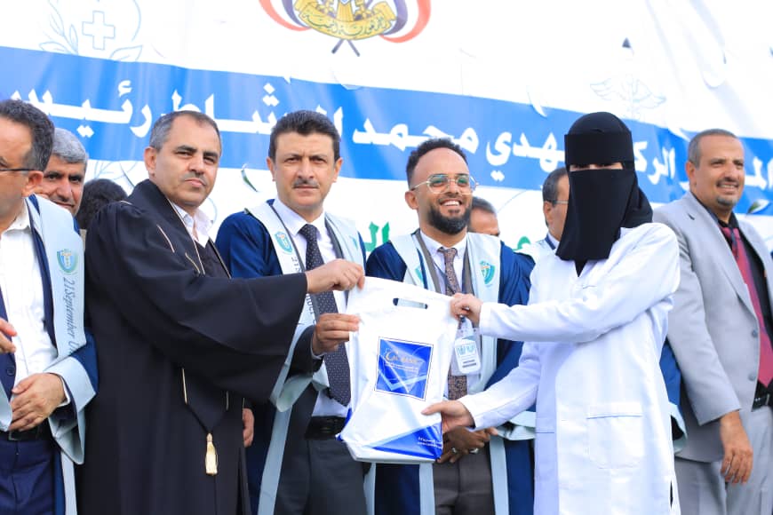 1003982221 - جامعة 21 سبتمبر Group of graduates and officials present a gift bag to a woman in a niqab at a ceremony, with a blue Arabic banner behind them.