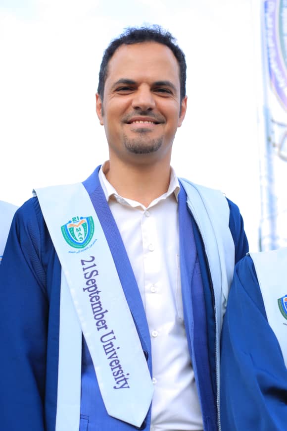 1003982224 - جامعة 21 سبتمبر Graduating student wearing a light-blue sash with the 21 September University logo over a white shirt and blue gown, smiling.
