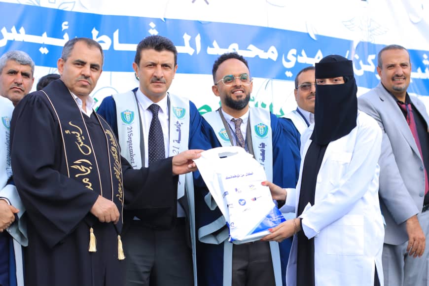 1003982227 - جامعة 21 سبتمبر Officials in ceremonial robes present a file bag to a woman in a black hijab and white lab coat during a graduation-style ceremony, with a blue banner in Arabic in the background.
