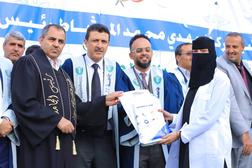 1003982230 - جامعة 21 سبتمبر Officials in ceremonial robes present a bag to a graduate during a formal ceremony with a blue banner in the background