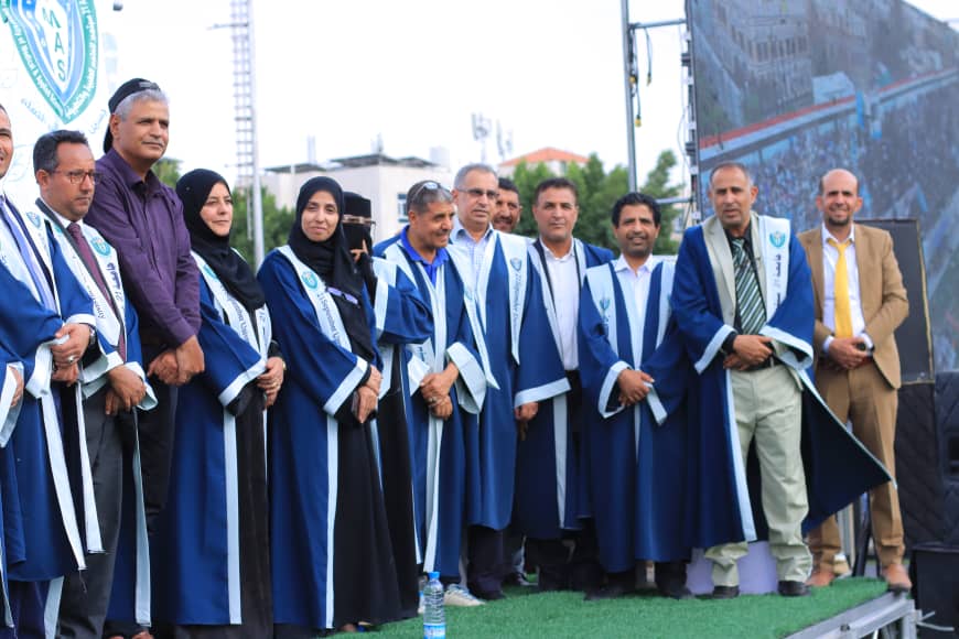 1003982233 - جامعة 21 سبتمبر Group of graduates in blue gowns standing on a stage outdoors, some wearing headscarves, posing for a photo.