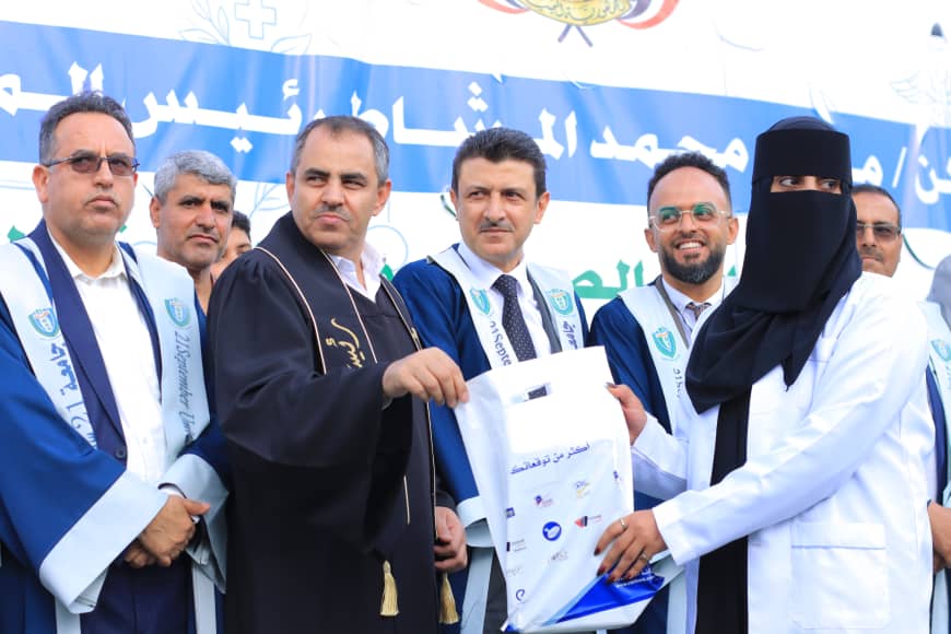 1003982236 - جامعة 21 سبتمبر Group of graduates in blue and white robes on stage while a man in a black robe presents a bag to a woman in a niqab at a ceremony, with officials watching.