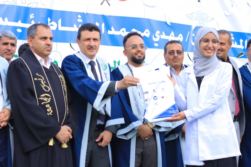1003982239 - جامعة 21 سبتمبر Group of professionals at a formal ceremony; a woman in a white coat hands a certificate to a man in blue academic robes, with a blue banner in the background.